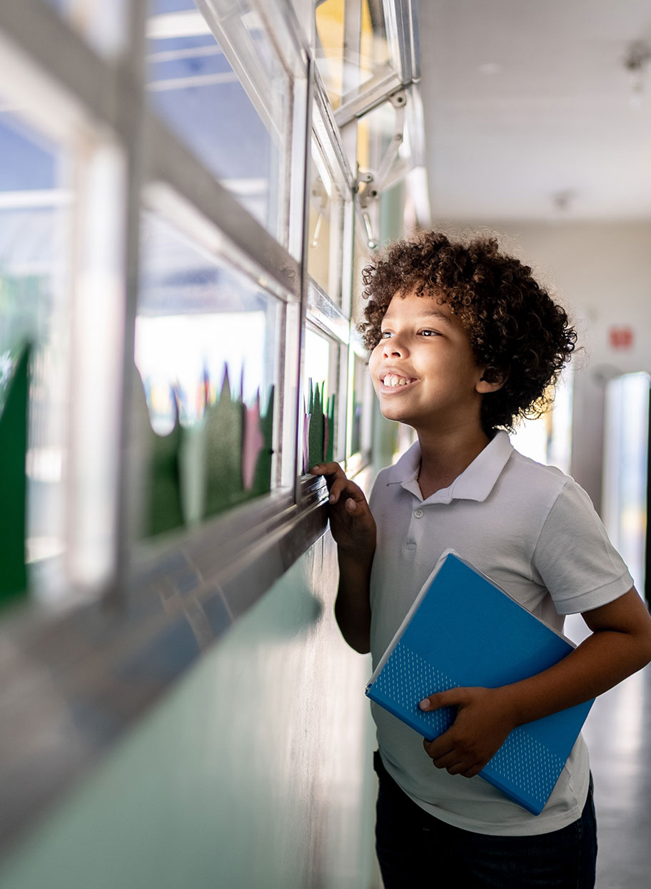 Young child holding a notebook looks out a school window